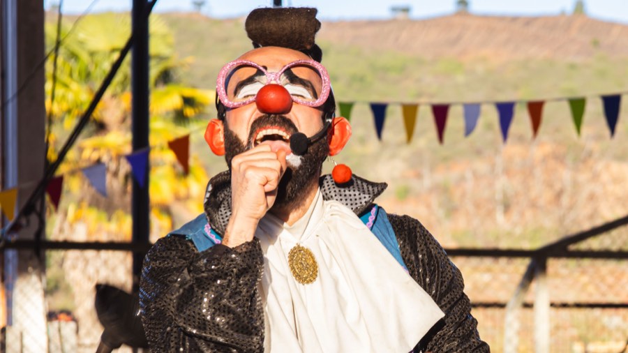 Payaso con nariz roja y anteojos actuando al aire libre bajo banderines de colores.